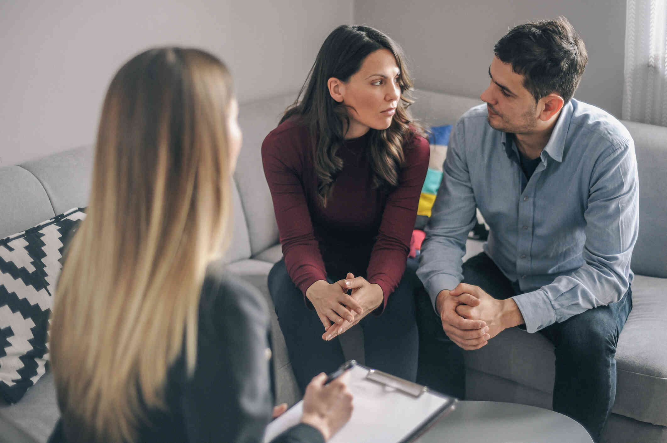 A male and female family member look at each other with serious expressions while sitting across from their female  family therapist.